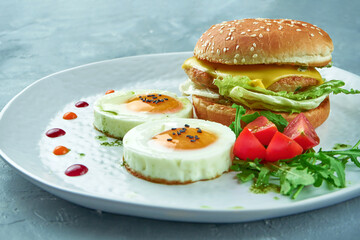 Perfect breakfast - chicken and cheese burger, 2 fried eggs and salad with arugula and cherry tomatoes in a white plate on a gray background