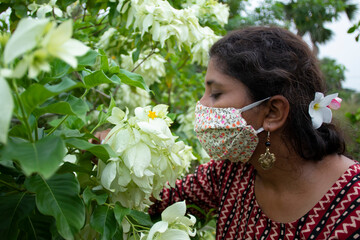 Beautiful lady with floral mask, earrings and designer top smells white Mussaenda flowers. Protection from corona in a stylish manner.
