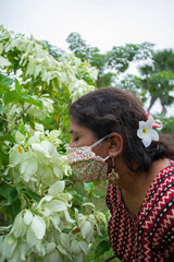 Beautiful lady with floral mask, earrings and designer top smells white Mussaenda flowers. Protection from corona in a stylish manner.
