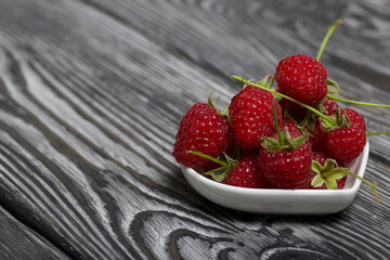 Raspberries with tails lie in a heart-shaped saucer. On black boards, with an expressive woody texture.