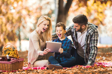 Happy family having picnic in autumn park, using digital tablet.