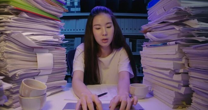 Young asian student woman is sitting at desk cover with stack of paperwork at home and looking at screen, typing on keyboard notebook. Alone teen girl using laptop studying hard overload at late night