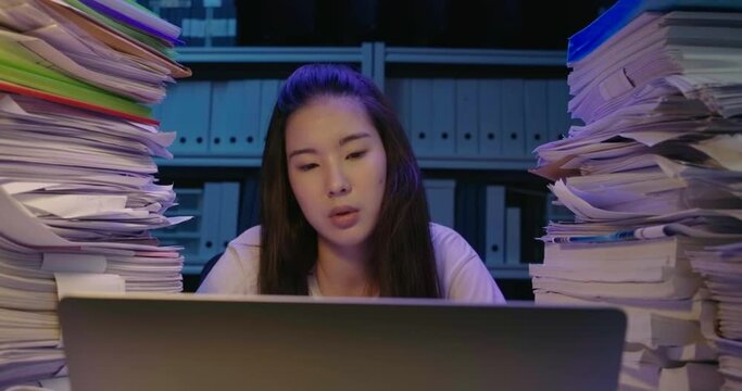 Young asian student woman is sitting at desk cover with stack of paperwork at home and looking at screen, typing on keyboard notebook. Alone teen girl using laptop studying hard overload at late night