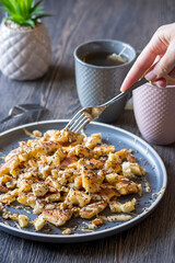 A hand grabbing with a fork a gluten-free, shredded pancake / kaiserschmarrn served with halvah spread, banana, walnuts, and grated chocolate placed on a decorative plate and teacups at the background