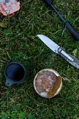 a tin can with stew, a mug with coffee, a fishing rod, a knife, a box of spoons lie on the grass.Top view