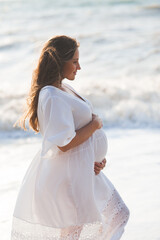 Fototapeta na wymiar Beautiful elegant pregnant woman 25-26 year old wearing white trendy dress holding tummy with hands posing over sea shore background close up. Motherhood. Maternity.