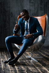 Black male businessman in a classic suit sits in an office chair, smiles and raises his thumb up