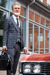 Fototapeta na wymiar handsome senior businessman near his car outdoors, stylish bearded male in elegant suit after business meeting, next to luxurious representative red auto