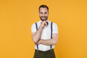 Smiling handsome young bearded man 20s wearing white shirt bow-tie suspender posing standing put hand prop up on chin looking camera isolated on bright yellow color wall background, studio portrait.
