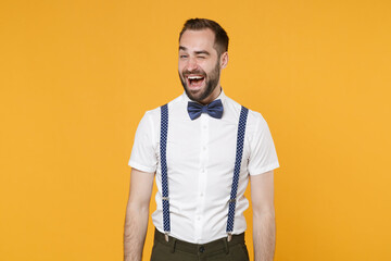 Smiling blinking cheerful funny young bearded man 20s wearing white shirt bow-tie suspender posing standing and looking camera isolated on bright yellow color wall background, studio portrait.