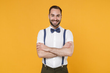 Smiling handsome attractive young bearded man 20s wearing white shirt bow-tie suspender posing holding hands crossed looking camera isolated on bright yellow color wall background, studio portrait.