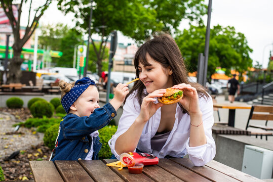 Young mother and little daughter ear burger together in outdoors cafe