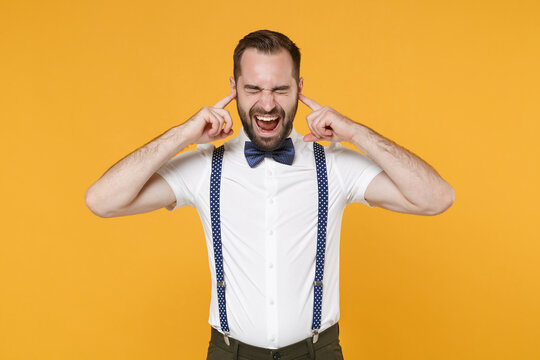 Crazy frustrated young bearded man 20s in white shirt bow-tie suspender posing covering ears with fingers screaming keeping eyes closed isolated on bright yellow color wall background studio portrait.