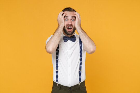 Shocked worried perplexed young bearded man 20s in white shirt bow-tie suspender posing standing put hands on head looking camera isolated on bright yellow color wall background studio portrait.