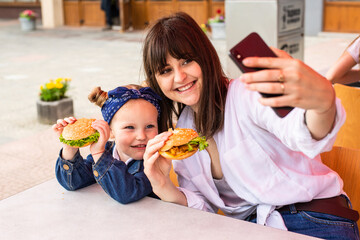 Obraz na płótnie Canvas Young mother with little girl eating a hamburger take selfie on the street cafe