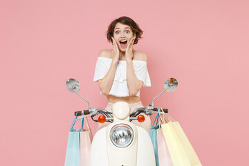 Surprised amazed young woman in white summer clothes hold package bag with purchases after shopping put hands on cheeks sitting driving moped isolated on pastel pink colour background studio portrait.