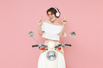Cheerful young woman 20s in white summer clothes hat glasses listening music with headphones dancing pointing index fingers up driving moped isolated on pastel pink colour background studio portrait.