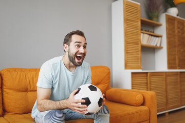 Excited young bearded man football fan 20s wearing basic blue t-shirt cheer up support favorite team with soccer ball keeping mouth open sitting on couch resting spending time in living room at home.