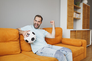 Joyful young bearded man football fan 20s in basic blue t-shirt cheer up support favorite team with soccer ball doing winner gesture sitting on couch resting spending time in living room at home.