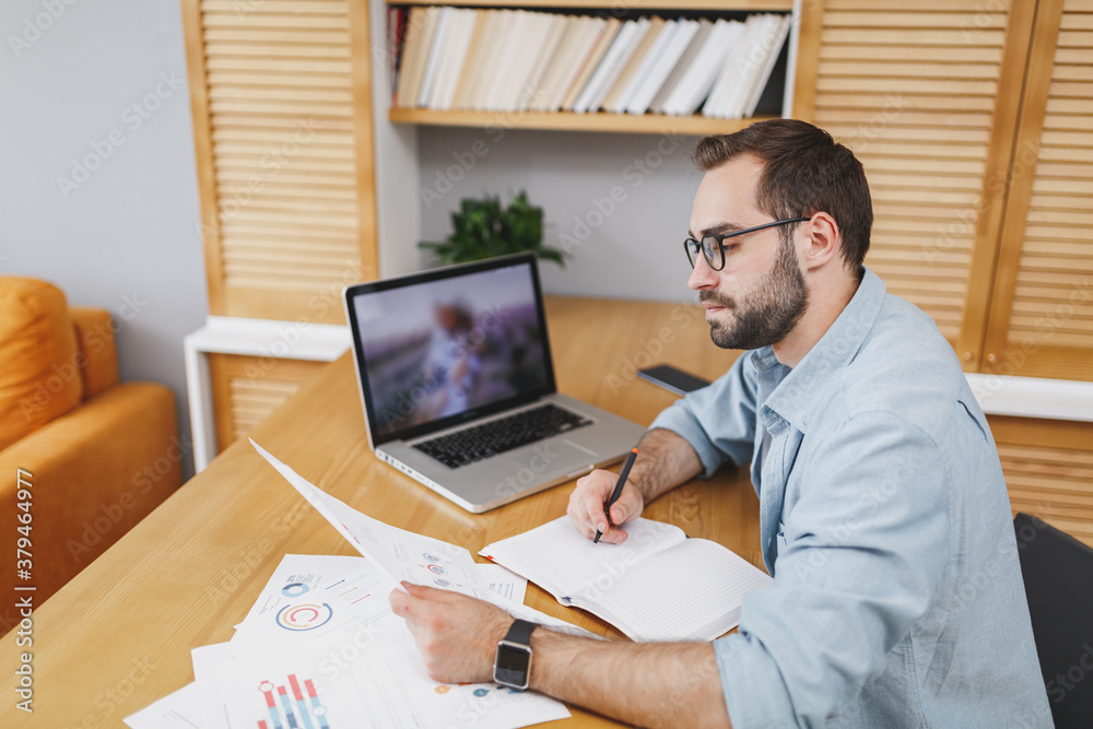 Wall mural handsome serious successful young bearded business man 20s wearing blue shirt glasses sitting at des