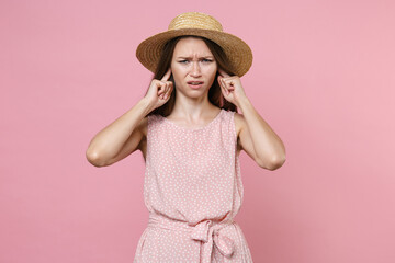 Displeased concerned puzzled young brunette woman 20s wearing pink summer dotted dress hat posing covering ears with fingers looking camera isolated on pastel pink color background studio portrait.