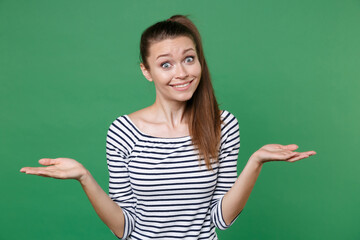 Confused smirked perplexed puzzled young brunette woman 20s wearing striped casual clothes posing standing spreading hands looking camera isolated on green color wall background studio portrait.