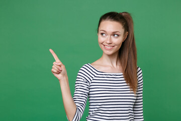Smiling attractive young brunette woman 20s wearing striped casual clothes posing standing pointing index finger aside up on mock up copy space isolated on green color wall background studio portrait.