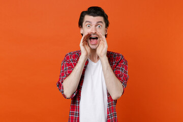 Shocked excited young brunet man 20s wearing white t-shirt red checkered shirt posing screaming with hands gesture near mouth looking camera isolated on orange color wall background studio portrait.