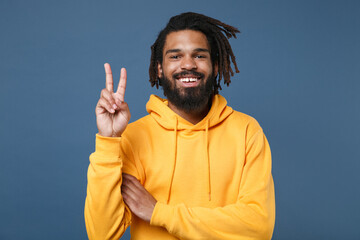 Smiling young african american man guy in casual yellow streetwear hoodie posing isolated on blue wall background studio portrait. People sincere emotions lifestyle concept. Showing victory sign.