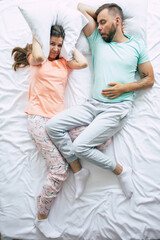 A stressed angry woman is covering her ears with help of a pillow due to snoring of her husband....
