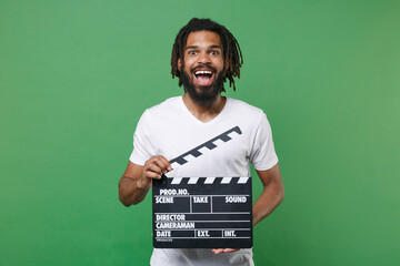 Surprised young african american man guy with dreadlocks 20s wearing white casual t-shirt posing holding classic black film making clapperboard isolated on green color wall background studio portrait.