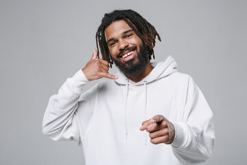 Smiling young african american man guy 20s in white casual streetwear hoodie doing phone gesture like says call me back point index finger on camera isolated on grey color background studio portrait.