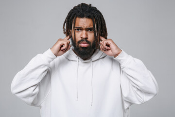 Upset young african american man guy with dreadlocks 20s wearing white casual streetwear hoodie posing looking camera covering ears with fingers isolated on grey color wall background studio portrait.