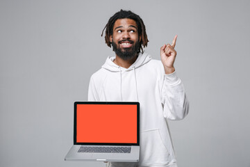 Smiling african american man 20s in white streetwear hoodie hold laptop pc computer with blank empty screen pointing index finger up on mock up copy space isolated on grey background studio portrait.