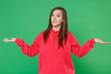 Confused puzzled concerned young brunette woman 20s wearing bright red casual streetwear hoodie posing spreading hands looking camera isolated on green color wall background studio portrait.