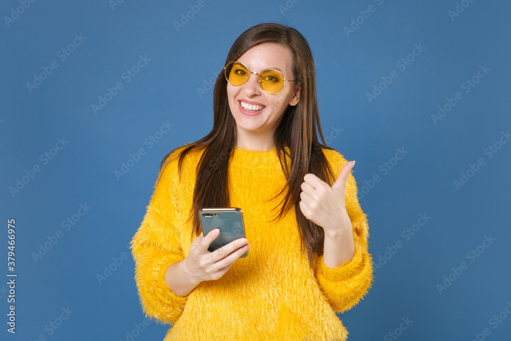 Sticker Smiling funny young brunette woman 20s wearing yellow fur sweater eyeglasses posing using mobile cell phone typing sms message showing thumb up isolated on blue color wall background studio portrait.