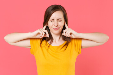 Fototapeta premium Displeased dissatisfied tired young brunette woman 20s wearing yellow casual t-shirt posing standing covering ears with fingers keeping eyes closed isolated on pink color background studio portrait.