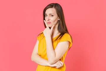 Pensive confused perplexed concerned young brunette woman 20s wearing yellow casual t-shirt posing standing put hand prop up on chin looking camera isolated on pink color background studio portrait.