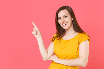 Cheerful smiling beautiful young brunette woman 20s wearing yellow casual t-shirt posing pointing index finger aside up on mock up copy space isolated on pink color wall background studio portrait.