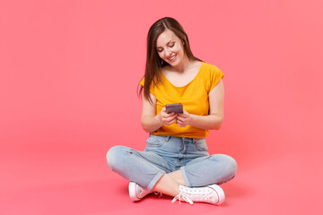 Obraz na płótnie Canvas Full length portrait of smiling beautiful attractive young brunette woman 20s in yellow t-shirt sit on floor using mobile cell phone typing sms message isolated on pink color wall background studio.