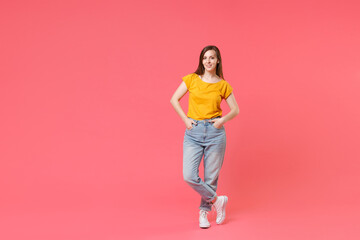 Fototapeta na wymiar Full length portrait of smiling beautiful young brunette woman 20s wearing yellow casual t-shirt posing standing holding hands in pockets looking camera isolated on pink color wall background studio.