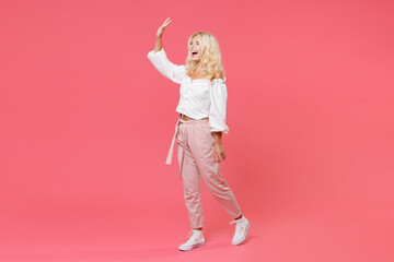 Full length portrait of cheerful young blonde woman 20s wearing white casual clothes waving and greeting with hand as notices someone looking aside isolated on bright pink colour background in studio.