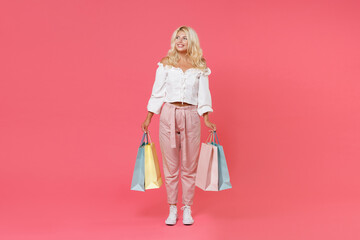 Full length portrait of smiling young blonde woman 20s in white casual clothes standing hold package bag with purchases after shopping looking aside isolated on bright pink colour background studio.