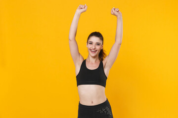 Happy cheerful young fitness sporty woman 20s wearing black sportswear posing training clenching fists doing winner gesture rising hands isolated on bright yellow color background studio portrait.