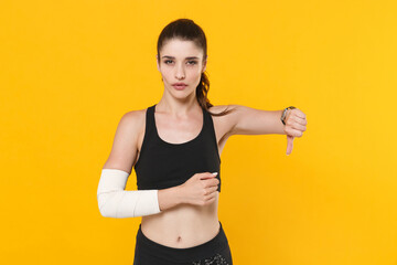 Dissatisfied young fitness sporty woman 20s wearing black sportswear posing training get injured arm elbow with elastic bandage showing thumb down isolated on bright yellow background studio portrait.