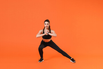 Fototapeta na wymiar Full length portrait of beautiful young fitness sporty woman in black sportswear posing training working out doing stretching exercise lunge looking camera isolated on orange color background studio.
