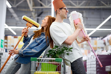 joyful married couple have fun in the market, young caucasian man and woman hold mops in hands, dance and sing
