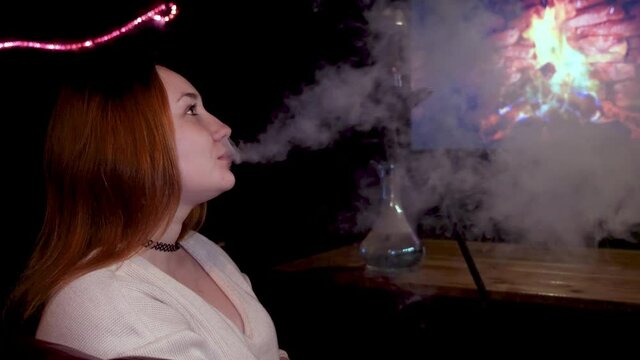 Side view of a red haired girl smoking shisha in front of TV screen with burning fire. Media. Young beautiful woman with a black choker around her neck smoking hookah on the background of fake