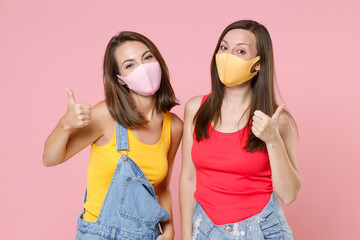 Two funny young brunette women friends in casual denim clothes sterile face mask to safe from coronavirus virus covid-19 showing thumbs up isolated on pastel pink colour background studio portrait.