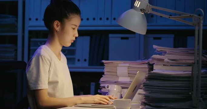 Young asian student woman is sitting at desk cover with stack of paperwork and typing on keyboard laptop. Alone teen girl stretch oneself for relax from stress while studying hard at late night.